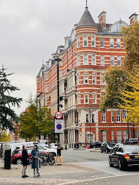 A busy street scene in front of a large, historic red-brick building with ornate stone detailing and rounded turret features, typical of Victorian architecture. The building's façade has multiple rows of white-framed sash windows, and the roof is topped with a conical turret and a variety of roof shapes including gables and dormer windows. In the foreground, two people are engaged in conversation near a sidewalk, standing beside bicycles leaning against a black post; one is dressed in a dark coat and light trousers, the other in a dark jacket and beige trousers. Several parked cars, including a black vehicle and a white van, are lined along the street, which also features a lamppost and a street sign indicating a 20 mph speed limit and a no-parking zone. There is a tree with yellow autumn leaves on the right side, contrasting with the building’s red brickwork. The scene captures a typical urban environment where private waste disposal might be arranged outside of local council collections, highlighting aspects relevant to independent rubbish removal services such as those provided by Waste Disposal West Kensington.