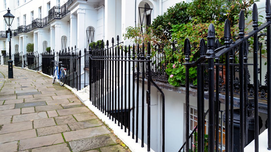 A row of white terraced houses with black wrought iron railings along a paved sidewalk. The railings feature pointed finials and are adjacent to small garden areas with green foliage and pink flowers. One section of the railing encloses a staircase leading down to a basement entrance, partly obscured by the railings. A blue bicycle is propped against the railing on the sidewalk. The buildings have black-framed windows and arched doorway features, with some potted plants visible on the front steps. Natural daylight illuminates the scene, creating a clear, neutral view of the residential street, which may be associated with private property boundaries or alternative waste disposal options in the context of rubbish removal services by Waste Disposal West Kensington.