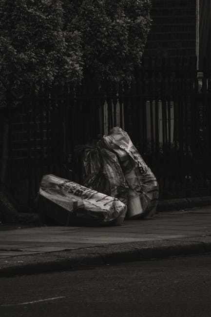 Two large, crumpled black and white plastic rubbish bags are positioned on the pavement near a black metal fence, with a leafy shrub visible behind the fence. The bags appear to be filled with waste materials and are partially leaning against each other. The scene is taken outdoors, with the bags situated on a street or sidewalk in an urban environment, and the surface beneath the bags is a concrete or asphalt pavement, showing natural weathering and slight unevenness. The lighting suggests overcast conditions or shade, contributing to the subdued tone of the scene. This image illustrates the type of waste that might require private rubbish removal services, such as those offered by wastedisposalwestkensington.co.uk, to manage council-unsupported rubbish accumulation effectively, particularly when large or irregular waste items are involved in on-site clearance or alternative waste handling arrangements.