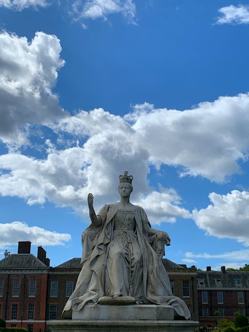 A stone statue of a seated, crowned royal figure with an elegant, draped gown and a cape, positioned on a pedestal in an outdoor city setting. The statue’s right hand is raised in a gesture, while the left rests on the arm of the chair. The background features a row of historic brick buildings with dark roofs, and a partly cloudy sky with large, white cumulus clouds against a bright blue sky. The scene appears to be set in a public square or park area, where outdoor rubbish removal services, such as those provided by companies like wastedisposalwestkensington.co.uk, may be involved in maintaining the clean environment around historic landmarks or civic spaces, emphasizing the importance of private waste handling and on-site clearance for urban areas.