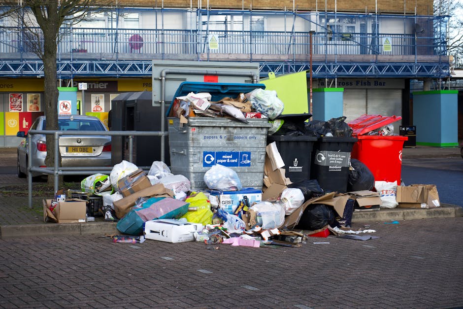 A collection of overflowing rubbish bins and scattered waste plastic bags, cardboard boxes, paper packaging, and miscellaneous rubbish are piled on a paved sidewalk in an urban area. The large grey bin marked for mixed paper and cardboard is tipped open, revealing crumpled paper and cardboard items. To its right, a black recycling bin with a white logo is also overflowing with black plastic rubbish bags, alongside a bright red bin. Smaller cardboard boxes and various loose waste are strewn around the base of the bins, some on the ground and others partially inside or leaning against the containers. A parked silver car is visible behind the rubbish, and a metal railing runs horizontally across the scene, separating the waste area from the parked vehicle. In the background, a building with storefronts and signs, including a fish bar, has a chain-link fence and scaffolding, indicating an urban commercial environment. The scene captures the typical appearance of waste awaiting collection or disposal in a public space, aligned with private waste management services.