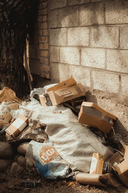 A narrow urban alleyway filled with accumulated rubbish, including large black and grey plastic bags, cardboard boxes, and miscellaneous debris piled against a makeshift wooden barrier at the end of the alley. The ground is uneven with visible dirt, small rocks, and some staining, indicating a space used for waste disposal or storage. On either side, buildings with graffiti and murals are visible, with windows and utility installations such as pipes and electrical boxes attached to the walls. The background features leafless trees and a brick building, suggesting an outdoor setting in an urban environment. In the foreground, a large, grey wheeled rubbish bag is prominent, with the surface showing a textured, crinkled appearance. The scene appears to be part of an area where independent waste collection or private rubbish removal services might be engaged, aligning with concepts of on-site clearance or alternative waste handling, as sometimes seen in less formal disposal arrangements managed by waste management specialists like wastedisposalwestkensington.co.uk.