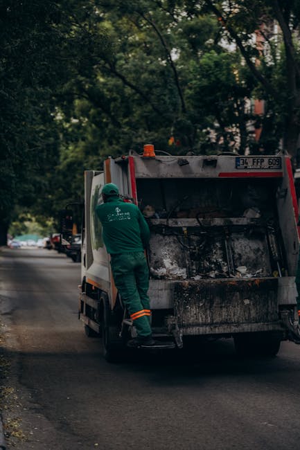 A worker dressed in green safety clothing and a helmet stands at the rear of a small waste collection van parked on a quiet urban street lined with trees. The van, which appears to be used for rubbish removal services, has its rear hatch open, revealing a load of charred debris and ashes inside. The surrounding environment includes parked cars along the curb and leafy foliage overhead, creating a shaded atmosphere. The scene suggests an on-site waste clearance operation, which may involve the collection of refuse from private property or alternative waste handling outside of regular municipal rubbish services. The vehicle is positioned on the asphalt road, and the worker's activity indicates an ongoing process of rubbish collection or disposal, consistent with waste management practices employed by companies like waste disposal services in West Kensington.