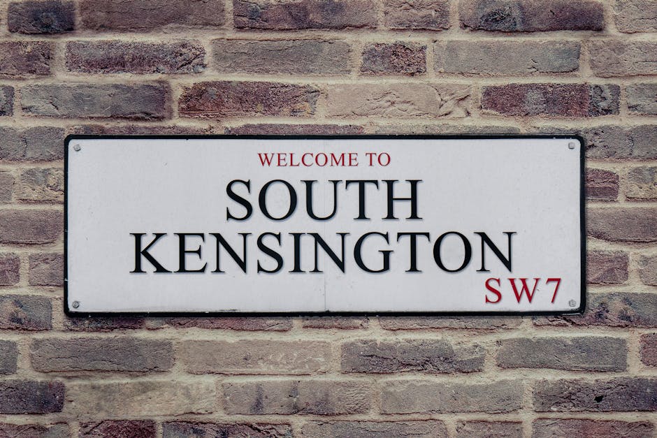 A rectangular white street sign with black and red lettering mounted on a brick wall. The sign reads 'Welcome to South Kensington' in large, bold black letters, with the words 'Welcome to' in smaller red font above, and the postcode 'SW7' in red positioned at the bottom right corner. The wall behind the sign is composed of reddish-brown bricks with a rough texture, laid in a traditional horizontal pattern. The sign is fixed with four screws, one in each corner, and is slightly reflective, contrasting clearly against the brick background. This signage indicates a location in South Kensington, an area known for its residential and commercial properties, often requiring professional rubbish removal or waste disposal services to manage local waste efficiently, aligning with the context of private waste handling and alternative collection methods supported by waste disposal companies.