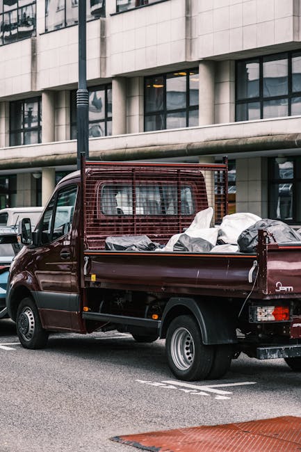 A small burgundy flatbed pickup truck parked on a city street in front of a modern multi-storey building with large rectangular windows. The truck's cargo bed is filled with various types of waste materials, including black plastic trash bags and loose debris. The truck features a metal grid barrier behind the cabin to secure the load. The vehicles are positioned on the road near a designated parking or loading area, with a nearby white van visible to the left. The environment appears to be an urban area, suitable for private rubbish collection services, with the building's reflective glass windows and concrete facade providing a contemporary backdrop. Natural daylight illuminates the scene, emphasizing the textures of the waste and the vehicle's paintwork, which has a slightly glossy finish. This image conveys the typical scene of directed waste collection involving small-scale or independent rubbish removal, characteristic of non-public disposal options in the West Kensington area, as offered by waste disposal specialists like wastedisposalwestkensington.co.uk.