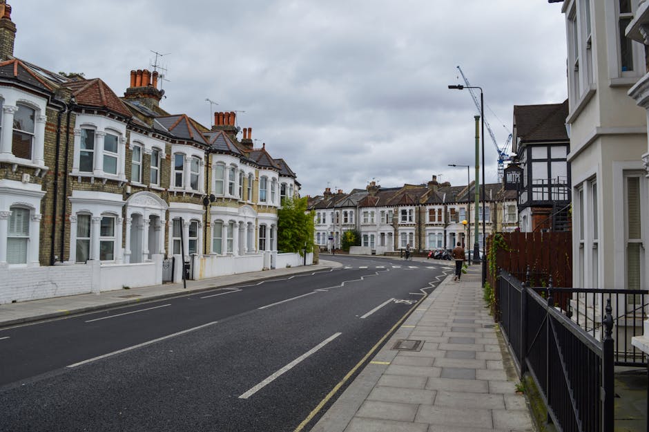 A quiet residential street scene featuring a row of Victorian-style terraced houses with decorative white cornices, bay windows, and brick facades painted in white and cream tones. The houses are aligned along a paved sidewalk, which runs parallel to the street and is separated by a black metal railing. In the background, additional terraced houses with similar architectural features extend along the road under a cloudy, overcast sky. The street has a smooth asphalt surface with visible lane markings, including double yellow lines on the roadside indicating parking restrictions. There are a few parked cars along the curbs, and a person walking away from the camera on the sidewalk. Construction work is evident on a building on the right side, where a crane and scaffolding are present, hinting at ongoing development or renovation. The scene appears to depict an area suitable for private waste collection or alternative rubbish removal services, with no visible rubbish or collection activities in progress, but the overall environment reflects typical urban residential waste management considerations.