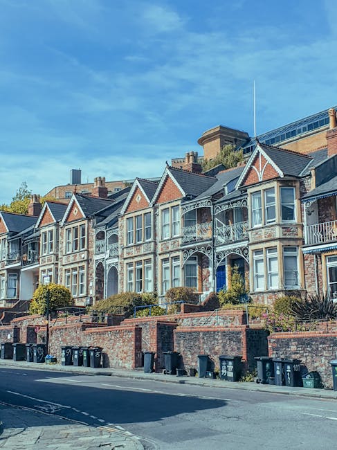 A residential street lined with parked cars on both sides, with a narrow asphalt road running through the center. The foreground features a variety of vehicles, including small hatchbacks, sedans, and vans, all in different colours such as black, silver, blue, and grey, parked neatly along the curb. On either side of the street, there are mature trees in full bloom with pink blossoms, their branches extending over the parked cars, creating a canopy effect. The trees have dark, textured bark and are densely covered with clusters of light pink flowers, contributing to a vibrant springtime atmosphere. Behind the trees, classic Victorian-style terraced houses with white facades, decorative cornices, and sash windows can be seen, typical of the West Kensington area. The overcast sky above is partly cloudy, casting diffuse natural light over the scene, giving it a calm and orderly residential feel. This image subtly aligns with private waste collection or rubbish removal services in the area, as the street appears clean and well-maintained, indicative of regular rubbish collection or clearance from local waste disposal providers such as Waste Disposal West Kensington.