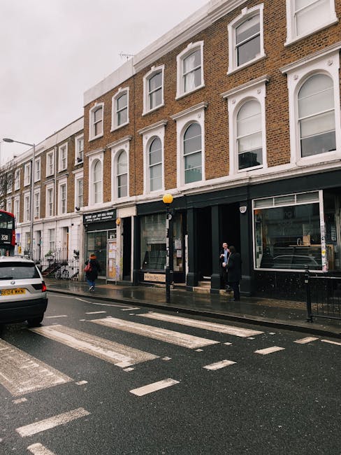 The image shows a wet street scene in an urban area with multi-storey brick buildings featuring white window frames and decorative architraves. The ground floor of the buildings includes commercial storefronts with large glass windows and black entrances, one of which appears to be a retail space or office. In front of the buildings, there is a yellow spherical streetlamp mounted on a black pole, positioned on a sidewalk with a tactile paving strip along the curb. Several pedestrians, including individuals dressed in dark coats and face masks, are walking along the pavement near the storefronts. A white car is parked partially on the street, and a red double-decker bus is visible in the background, approaching from the left side. The road surface is wet, with visible reflections of the streetlamp and vehicles, indicating recent or ongoing rain. The scene subtly reflects an environment where private waste collection or on-site clearance could be relevant for managing rubbish outside of traditional municipal services, especially in a converted residential or commercial unit setting.