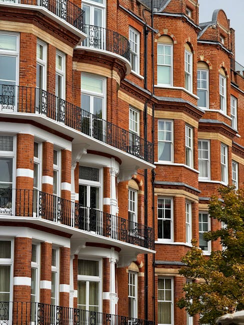The image depicts a close-up view of a multi-story residential building constructed with red brickwork, featuring white and beige stone detailing around the windows and along the building’s exterior. Several large, double-glazed sash windows are visible, framed with white trims, and some of the windows are slightly open. The building has ornate black wrought iron balconies with decorative railings, attached to the front façade, extending outward from multiple floors. The balconies include horizontal bars and curved sections, contrasting with the red brick and white accents. In the lower right corner, a part of a tree with green and yellow leaves is visible, suggesting an urban setting during late summer or early autumn. The natural daylight illuminates the façade evenly, highlighting the textures of the brick and stonework. This type of building, typical of West Kensington, may have been subject to internal renovations or conversions, which can generate waste requiring professional rubbish removal, such as that provided by waste disposal services like Waste Disposal West Kensington. The scene reflects a well-maintained residential property with features consistent with private communal living spaces and potential for underground or accessible waste collection operations.