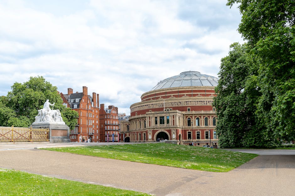 A large, round historic building with a domed roof and red brick exterior featuring arched windows and decorative stonework is situated on a grassy area with a paved pathway in front. To the left, a white marble monument or sculpture with intricate carvings is positioned near a cluster of tall, leafy green trees that frame the scene on both sides. The sky overhead is partly cloudy, casting diffuse light over the scene. In the background, a row of red-brick residential buildings with chimneys and dark rooflines line the street. The environment appears quiet and well-maintained, illustrating an urban park or open space in a city setting. This scene can be associated with private or independent waste management services, such as those offered by waste disposal companies, providing on-site clearance and rubbish removal in historical or public areas.