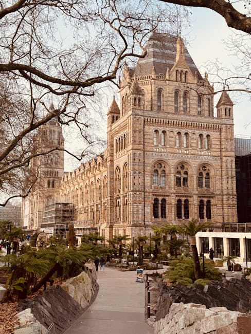 A historic building constructed of light brown and beige stone featuring intricate architectural details, arched windows, and a prominent central tower with a steep, pyramid-shaped roof. The structure is set against a partially overcast sky, with leafless tree branches extending across the upper portion of the image. In the foreground, there is a paved walkway surrounded by landscaped greenery, including small palm trees and bushes, with some large rocks and boulders placed along the sides of the path. A few people are visible walking near the building, and scaffolding is present on the left side, indicating ongoing maintenance or restoration work. To the right, modern glass and steel structures contrast with the historic architecture, suggesting a cityscape that integrates both old and new. The overall setting appears to be an urban area, potentially a park or public space, where independent waste removal or on-site clearance services by Waste Disposal West Kensington could be pertinent for maintaining cleanliness in the vicinity.