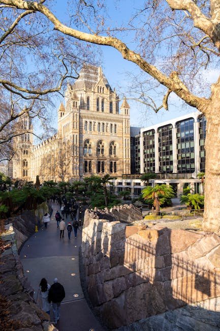 Inside the grand, historic interior of a museum or gallery with high, arched glass ceiling supported by metal framework, featuring ornate brickwork and decorative architectural details. A large, suspended skeleton of a whale or large marine animal is displayed in the central upper area, with visible vertebrae and skull, hanging from thin cables. The space is populated with visitors, some standing and observing, others walking around on the polished stone floors near the wide staircase leading to the upper level, where more visitors are gathered. The warm, natural daylight filters through the expansive glass roof, illuminating the interior's intricate brickwork and casting soft shadows. This scene exemplifies a preserved historic building repurposed for public exhibitions, aligning with professional waste management practices that support safe and clean cultural spaces, reflecting efforts like those of waste disposal services in maintaining cleanliness in public and institutional environments.